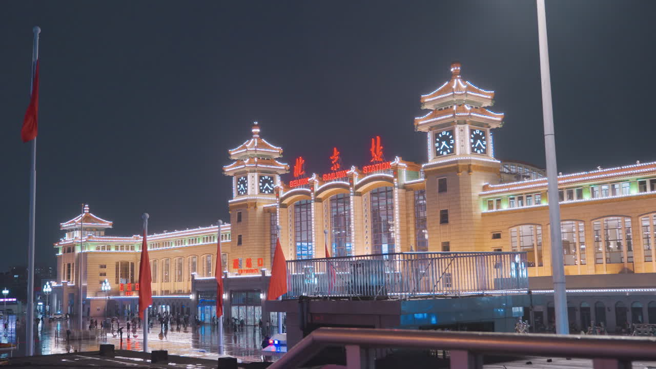 Geceleri canlı bir şekilde aydınlatılmış Çin istasyonu, kırmızı vurgularla aydınlatılmış görkemli cephe, yolcular ve turistlerle dolu hareketli bir meydan, dalgalanan bayraklar, güvenlik devriyesi ve satıcılar, platformun ötesinde varan trenler.