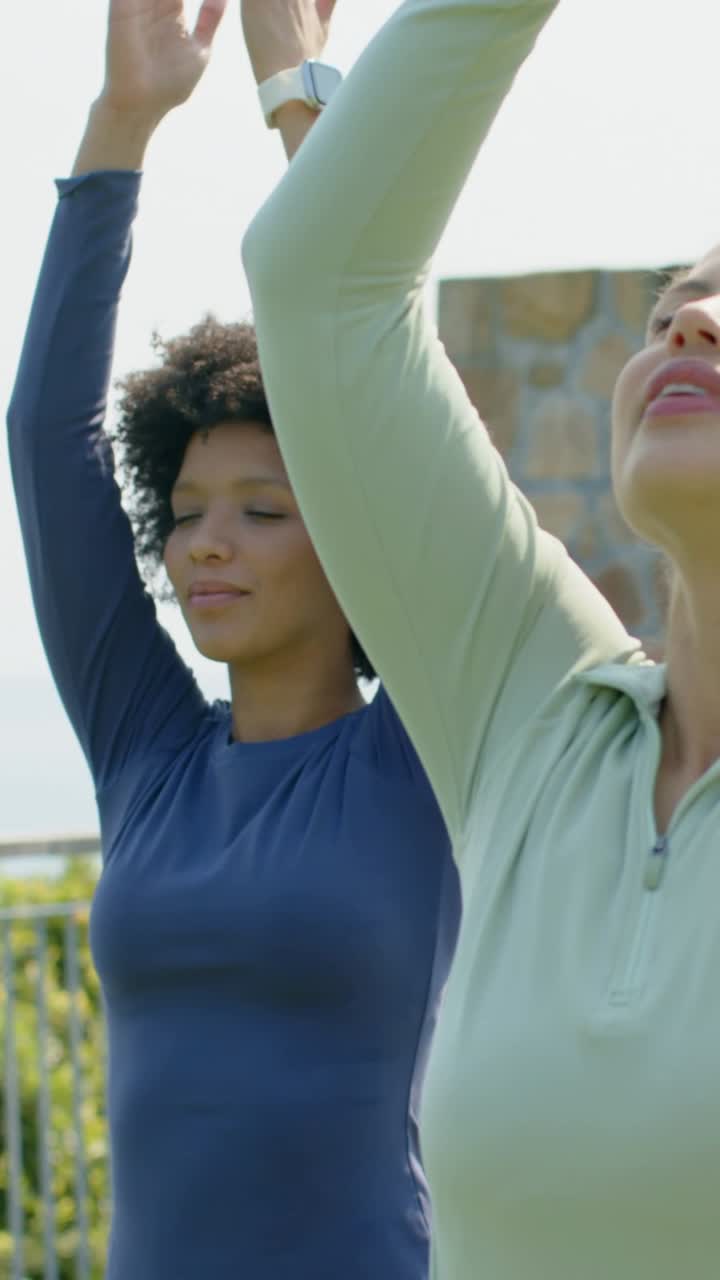 Vídeo vertical: Mulher de blusa azul-marinho, seguindo orientação, levantando os braços acima da cabeça para yoga em terraço com vista para o mar, com guarda-corpo.