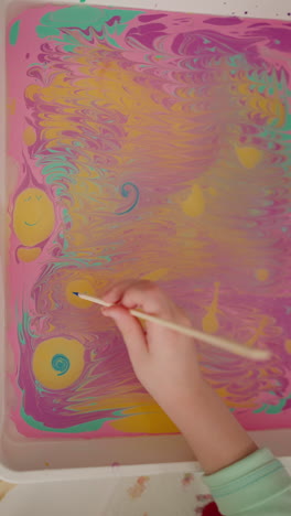 little student draws smiling face on oily water surface with paint in classroom close upper view slow motion. ebru design lesson for junior children