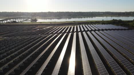 Vista-Aérea-Volando-A-Través-De-Filas-De-Paneles-Solares-Fotovoltaicos-Junto-Al-Río-Rural-Al-Atardecer