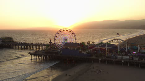 Pacific-Park-Theme-Park-on-Santa-Monica-Pier-with-Ferris-Wheel-in-beautiful-Golden-Hour-Sunset-light-over-coastline,-Aerial-Wide-Angle-Shot,-Lowering-crane-down