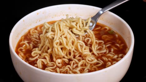 a metal fork lifts cooked instant noodles from a white bowl filled with steaming broth, shot in close-up with even studio lighting and a black background