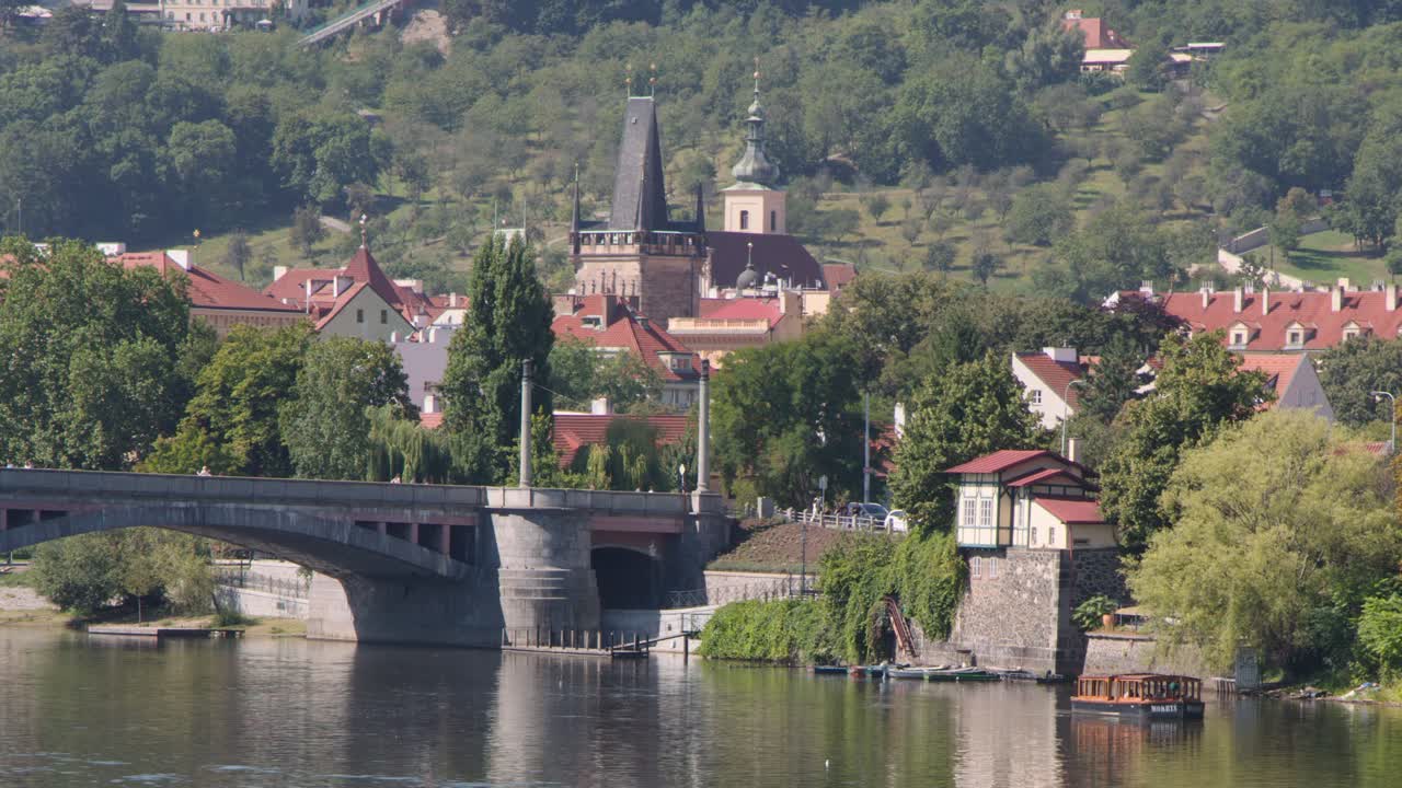 Steady daytime shot of Prague bridge, river, historic buildings, lush greenery, and cityscape
