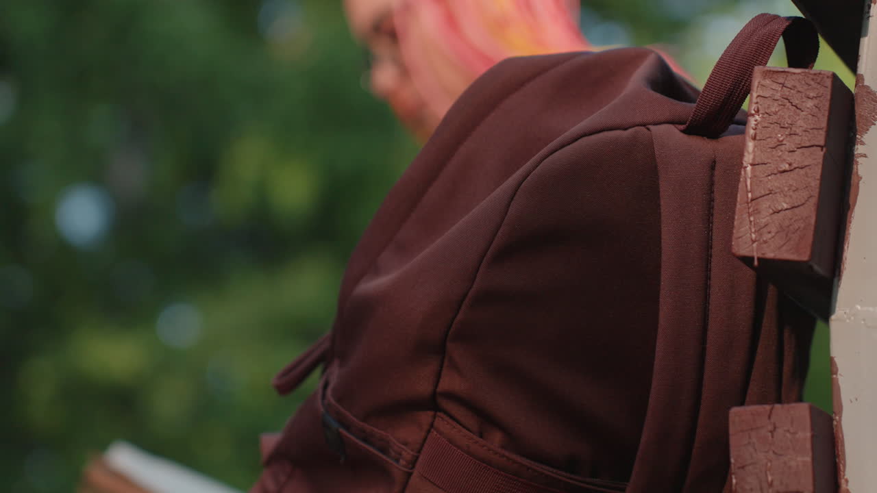 공원에 있는 벤치에 갈색 백팩이 놓여 있고 근처에 독자가 앉아 있어요. 흐릿한 녹색 배경에 가방끈과 주머니 디테일이 보입니다. 조용한 여행이나 공부 중 휴식을 암시하며 손을 뻗고 부드럽게 표현되었네요.