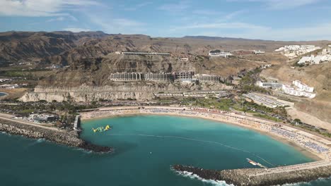 aerial backwards shot of beautiful bay with sandy beach and resort on hill during sunny day - playa de amadores, gran canaria