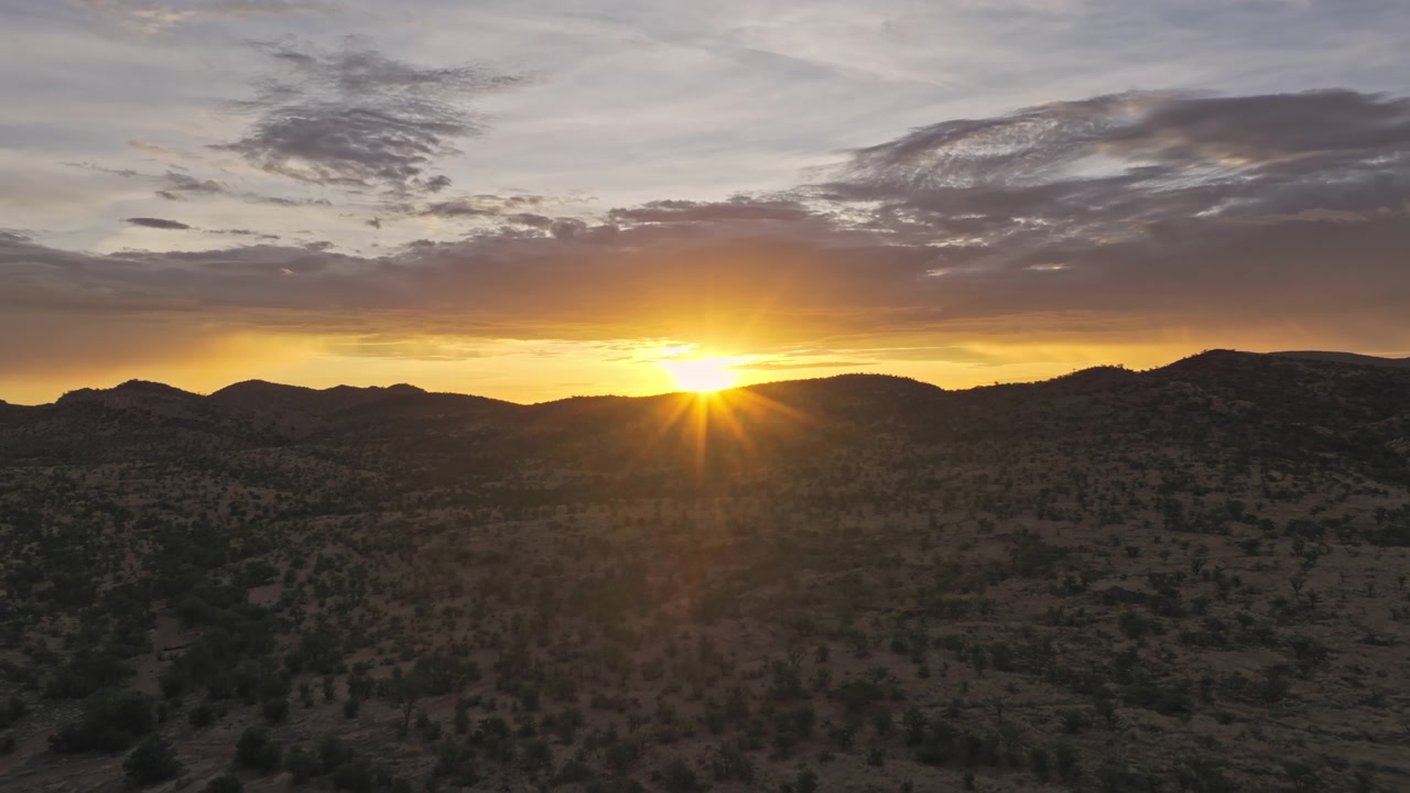 Vista aérea de um brilho solar intenso sobre colinas desérticas ondulantes na Namíbia central, com luz dourada espalhando-se por arbustos dispersos e terreno seco.