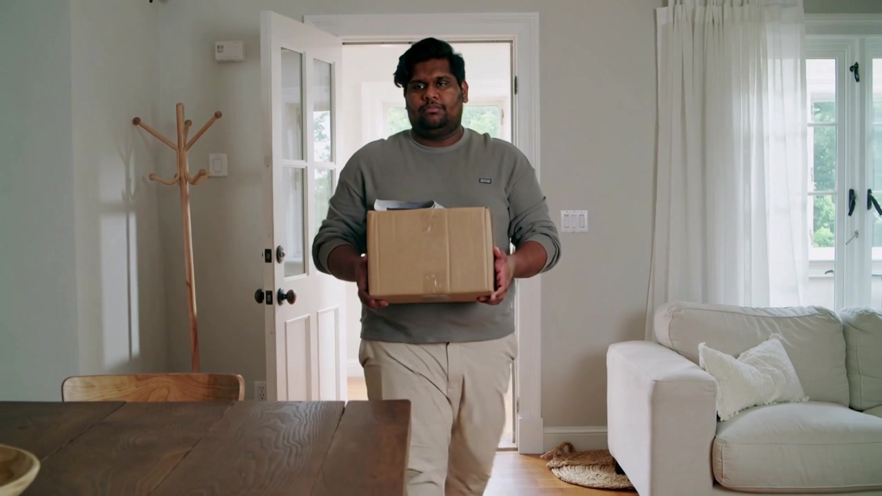 Door opening, man in gray sweater khaki pants bringing sealed box inside to dining table, paperwork
