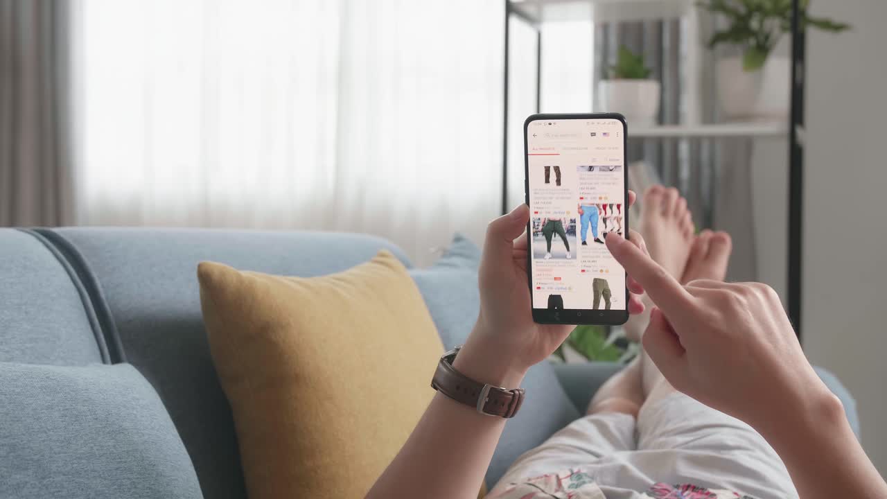 Close Up Of Hand Man At Home Lying On Couch In Living Room Using Smartphone Buys In Internet Shop. Man Looks At Goods In Online Store Clothing. Buy Fashion Clothes Directly On Smartphone