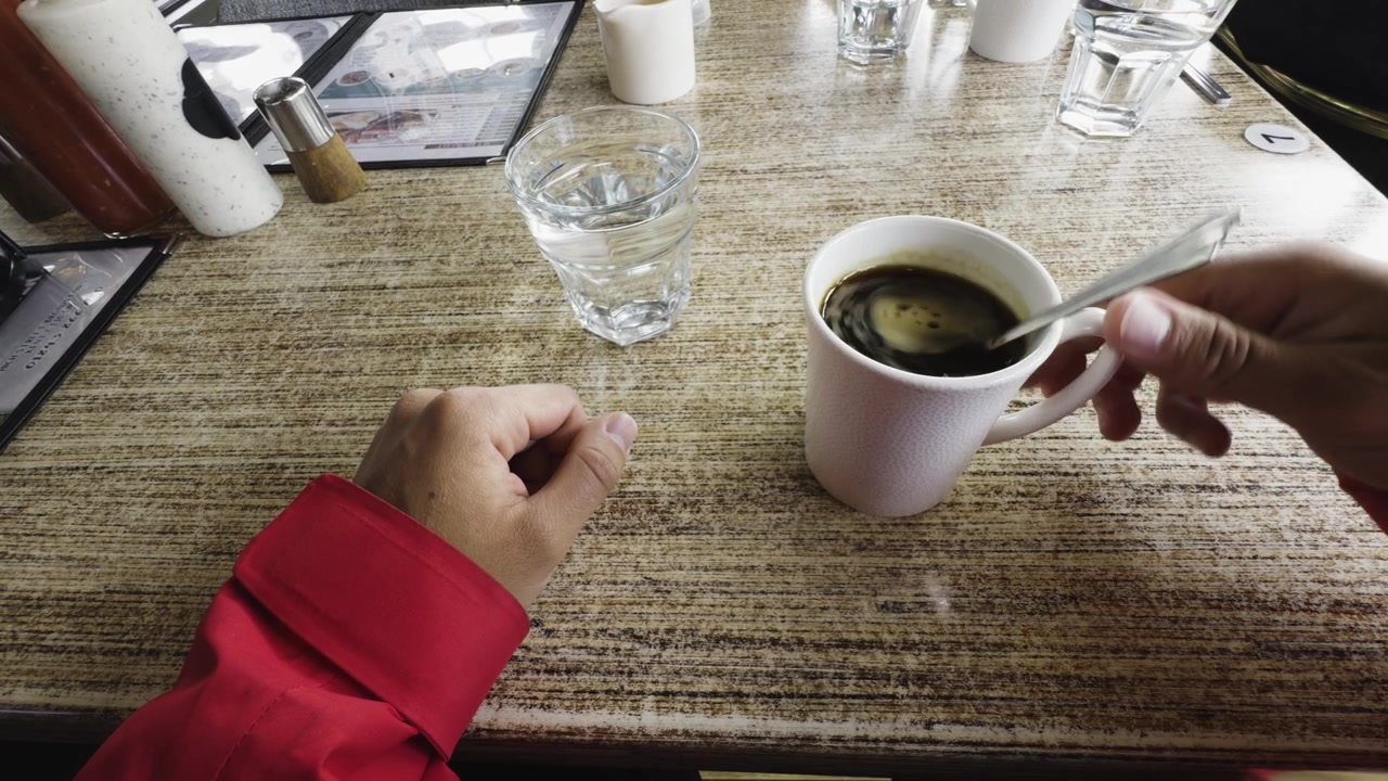 First-person POV of an individual in a red jacket sitting at a textured cafe table, stirring a hot cup of dark coffee with a spoon. A glass of water sits nearby. The mood feels warm and very calming.