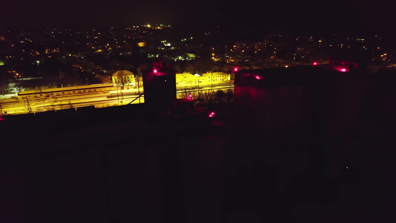 Dark silhouette of a silo marked with red lights in the industrial part of the city, with the background of an illuminated train station. Svitavy, Czech Republic.