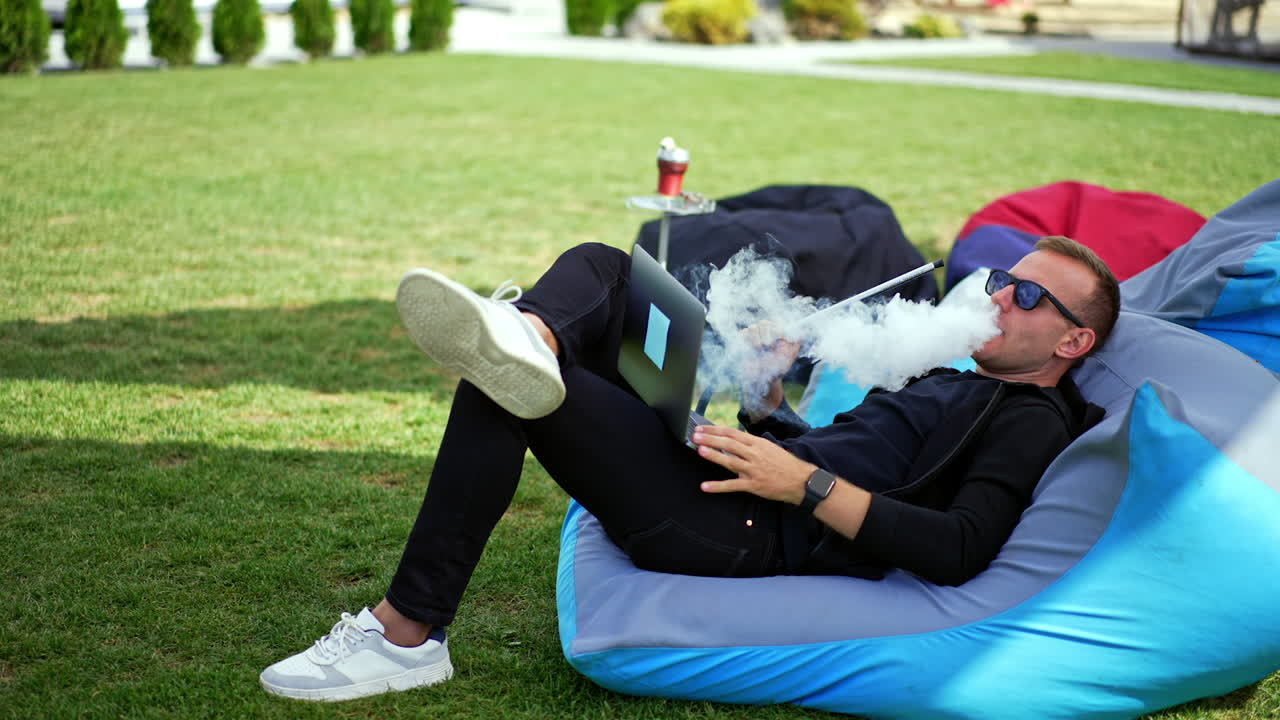 Mid-aged man in black clothes and sunglasses lies in bean bag chair outdoors. Resting male smoking hookah and looking at his laptop screen.
