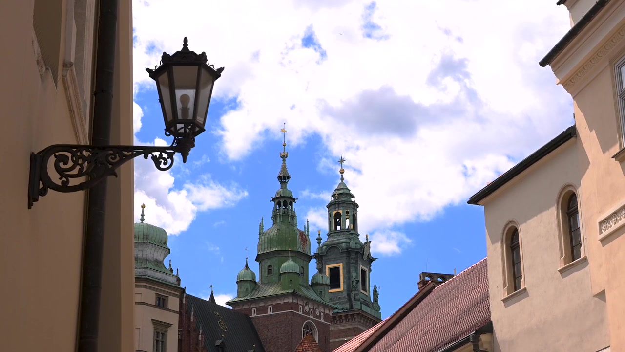 Dekorativ gatelykt og omkringliggende bygninger rammer inn Wawel-katedralens grønne kobbertårn under en lys, skyet himmel