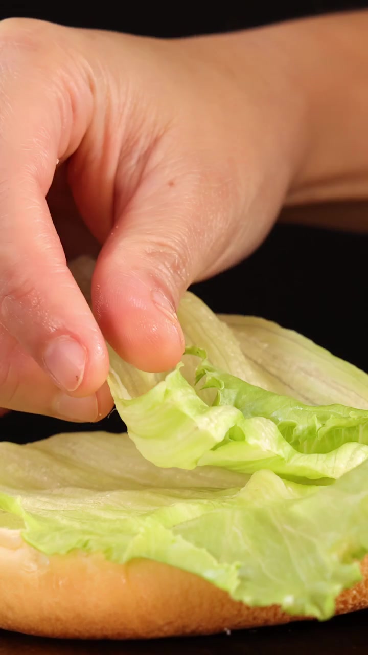 Una mano sistema delicatamente una fresca foglia di lattuga iceberg su un semplice panino da hamburger, ripreso in primo piano con illuminazione da studio brillante e uniforme e uno sfondo nero