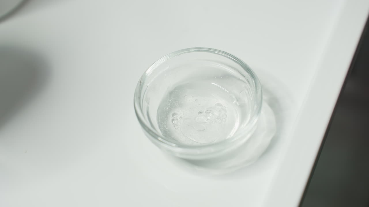 Transparent Bowl Placed On White Tray As Clear Viscous Gel Drips Drop By Drop From Container Tip Under Gentle Spa Lighting Close Up Of Skincare Prep Ritual Showcasing Delicate Texture And Purity