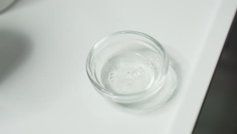 transparent bowl placed on white tray as clear viscous gel drips drop by drop from container tip under gentle spa lighting close up of skincare prep ritual showcasing delicate texture and purity