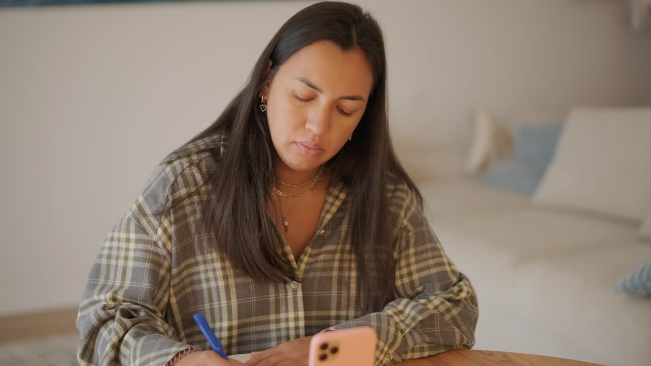A pregnant woman sitting at a wooden table in a cozy, softly lit home interior, writing or journaling. The scene exudes calm, focus, and a sense of preparation during maternity