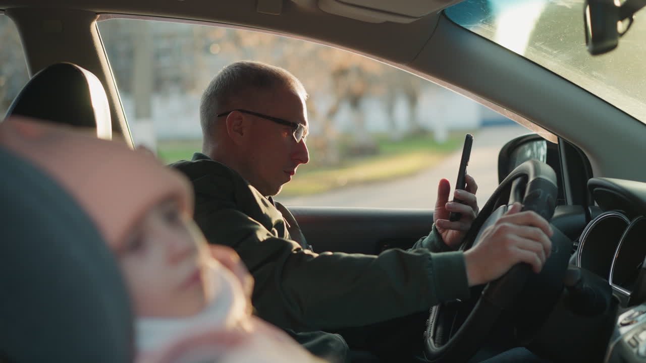 un hombre con una chaqueta verde está sentado en un coche, sosteniendo un teléfono en una mano y el volante con la otra. con una vista borrosa de una niña en el fondo