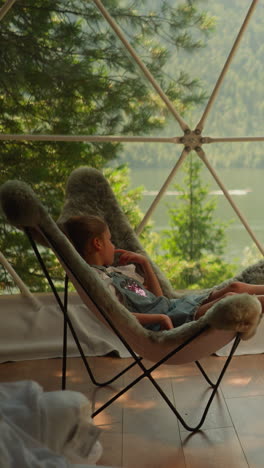 children in soft chairs under glamping dome. barefoot boy and girl sit in fluffy armchairs near panoramic window. little kids rest at camp on holiday