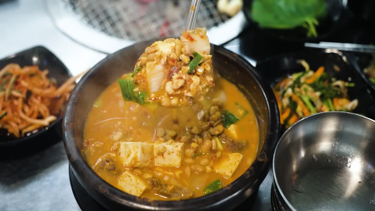 A ladle scooping hot, bubbling Korean Doenjang Jjigae soybean paste stew with tofu, vegetables, and beef served in a black dolsot bowl on a restaurant table