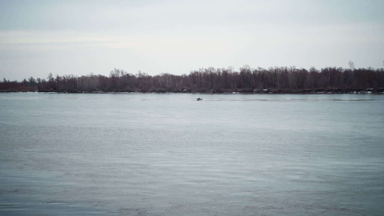 Wide view of calm river with small boat in distance near forested shoreline under cloudy sky during cold season, capturing peaceful water surface, natural landscape, outdoor environment
