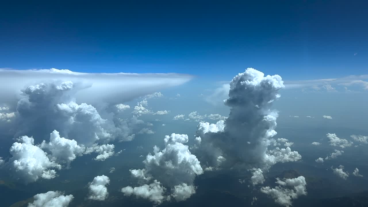 An immersive pilot’s view taken from a jet cockpit while flying above storm clouds under an intense blue sky, with an anvil shape cumulonimbus on the left.
