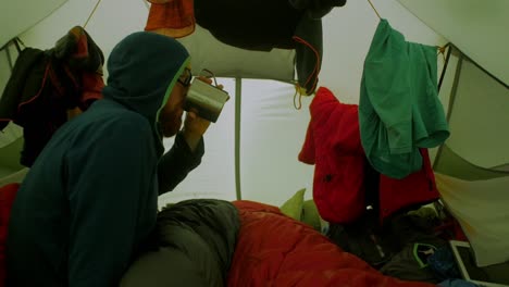 inside the tent during a stormy weather, male hiker in a sleeping bag waiting and drinking a beverage, tent walls are shaking in a gusts of wind, wide shot