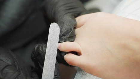 technician wearing black gloves gently files toenails of client resting leg on cushioned bed, focusing on hygiene and detail in professional pedicure session under bright light in clean environment