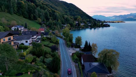 Luftaufnahme-Einer-Kurvenreichen-Zweispurigen-Asphaltstraße,-Die-Durch-Einen-Dichten-Grünen-Wald-Zu-Einem-Glitzernden-Blauen-See-Führt,-Mit-Schneebedeckten-Bergen-In-Der-Ferne