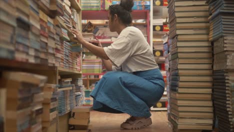 Asian-girl-sitting-and-exploring-books-through-bookshelves,-Side-angle-shot