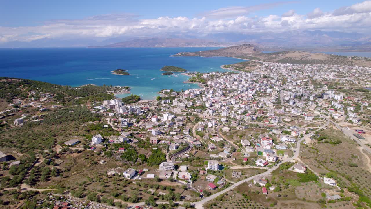Yüksek irtifalı drone görüntüsü, küçük adaların bulunduğu parlak turkuaz bir koyun yanında, kısmen bulutlu bir gökyüzü altında tepeler ve tarım arazileriyle çevrili, yoğun bir sahil kasabasını gösteriyor, akıcı kaydırma kamera hareketi