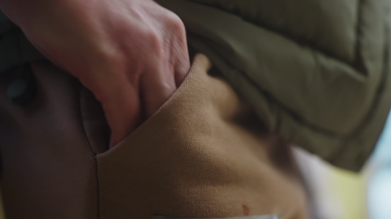 Closeup of human hand resting in brown pocket of casual pants, partially covered by green winter jacket, showing relaxed posture and natural gesture during quiet indoor moment in cool weather setting