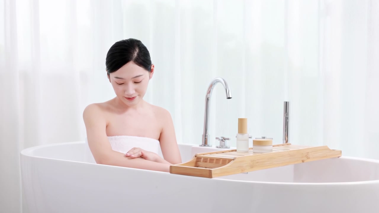 シャワー後のバスルームでバスタブに座って朝を過ごし、タオルに包まれた魅力的でゴージャスな女性が、栄養クリームやボディローションを塗って日常のスキンケアを行っている。スキンケア、セルフラブ、ボディケア、スパ。