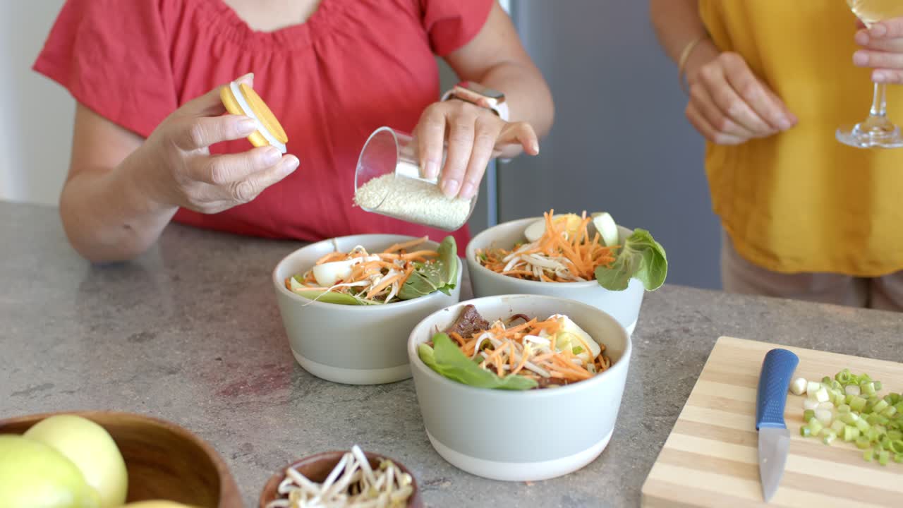 Senior and mature women seasoning three salad bowls with seed jar at kitchen counter after plating