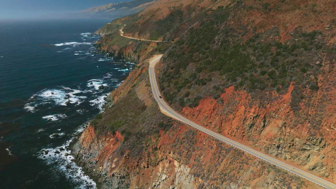 Magnificent mountains of California with highway in the middle. Dark blue ocean with white foamy waves. Aerial perspective.