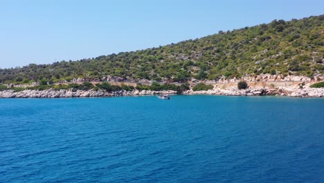 Amplia-Vista-Aérea-De-Una-Colina-Verde-Junto-Al-Mar-Mediterráneo-Azul-Tropical-En-Finike-Turquía-Con-Un-Velero-Anclado-En-El-Agua