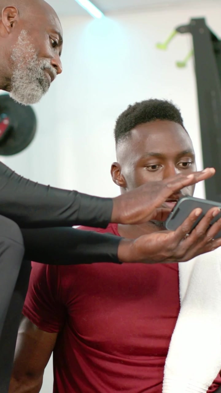Vertical video: Leaning coach reaching tapping phone showing client in maroon on gym bench coaching