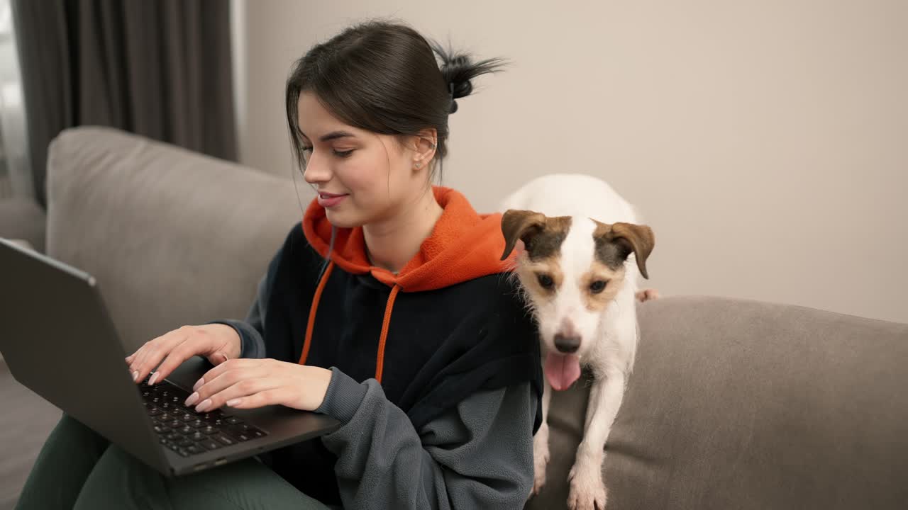 Young woman sitting on a couch working on laptop with her dog next her