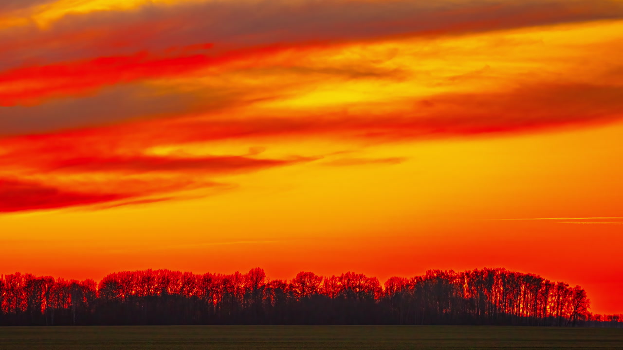 Dramatic telephoto time-lapse captures blazing diagonal cloud bands racing across fiery red and orange sky above dark silhouetted treeline and flat field in Latvian countryside