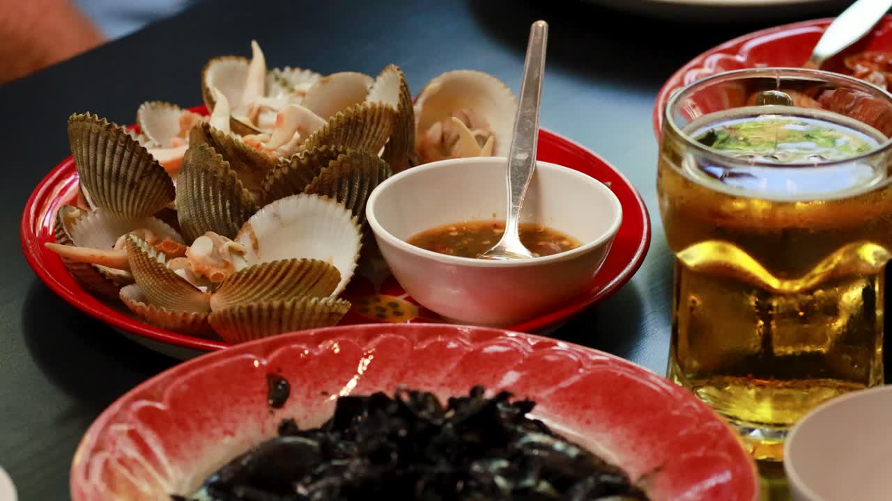 A vibrant seafood meal featuring clams, dipping sauce, and beer. Captured in a lively setting with warm lighting