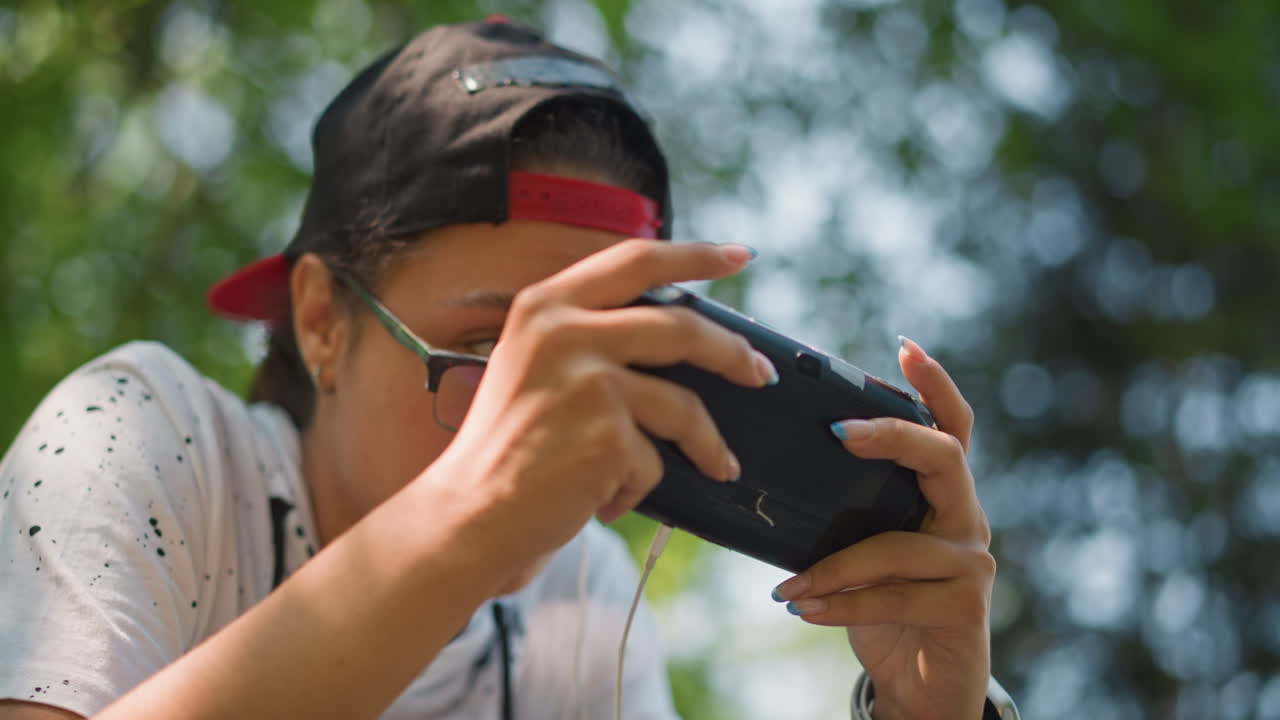 ragazzo asiatico adolescente che gioca intensamente in un parco, primi piani mostrano un giocatore con un berretto che stringe la cover del telefono, auricolari inseriti, occhiali scintillanti, alberi illuminati dal sole e sfondo bokeh, espressione concentrata del giocatore