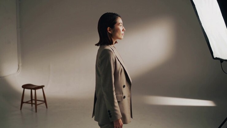 Alzando il mento, modella asiatica in blazer color cammello che regola la posa mentre la luce del softbox cambia, in posa per un ritratto