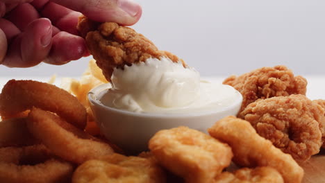 hand dipping fried chicken in creamy sauce with french fries and onion rings