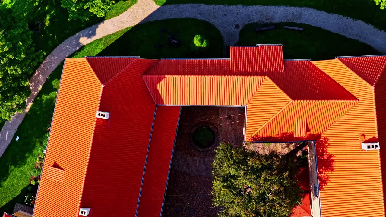 Colorful drone above Jaunpils Castle rooftops framed by green trees and morning light, medium top shot backdrop
