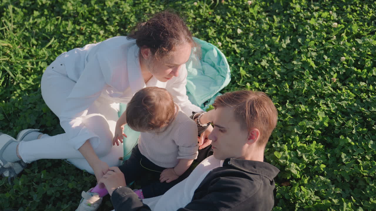 Weiße Eltern spielen mit ihrem Kleinkind; der Vater kitzelt das Kinn des Kindes, die Mutter richtet einen Schuh. Eine Picknickdecke ist in der Nähe, Sonnenlicht und Klee. Ungestelltes Lachen, spielerische Elternenergie und das Thema eheliche Fürsorge.
