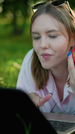 primer plano de una joven comiendo una manzana mientras está acostada en un campo cubierto de hierba, trabajando en una computadora portátil, disfrutando de un momento pacífico con la luz del sol filtrándose a través de los árboles