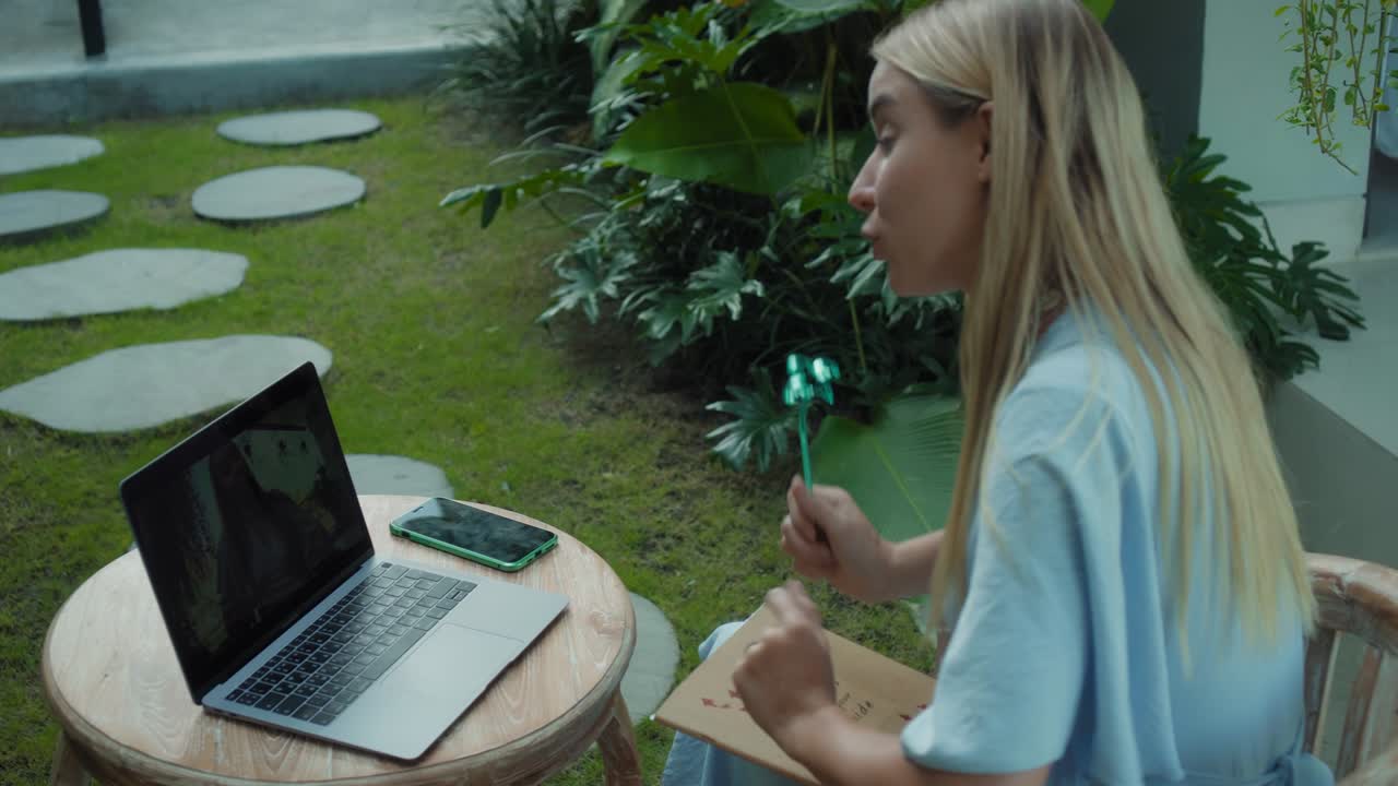 A girl in a blue dress remotely online working behind a laptop and looking into the screen the backyard with green plants