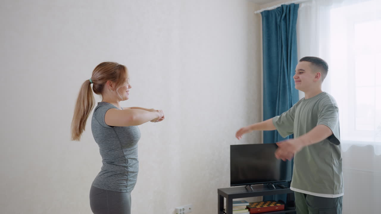 트레이너가 커튼이 드리워진 창문 배경 근처에서 웃는 얼굴들과 함께 가정 운동 세션 동안 팔 스트레칭을 하는 소년을 지도하며 격려, 지지, 피트니스, 건강, 자세 인식 등을 보여주고 있어요.