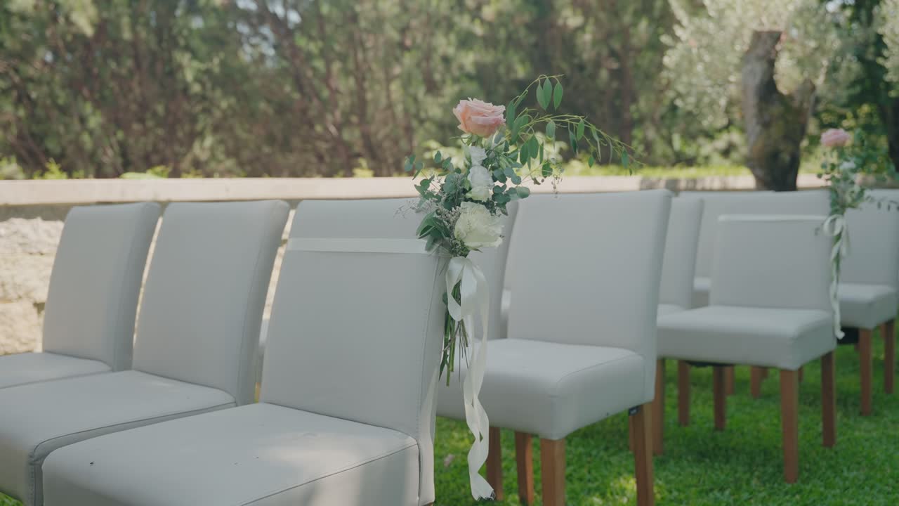 Rows of white chairs arranged on green grass with floral bouquets and ribbons attached for an outdoor wedding setup