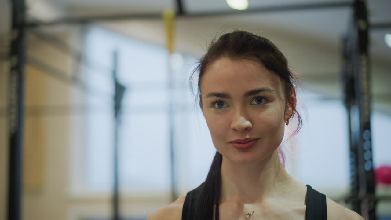 Ritratto di atleta donna sorridente in palestra, bagliore post-allenamento con capelli umidi e luce che accarezza la pelle, atmosfera di briefing con l'allenatore e pausa idratazione, sguardo amichevole verso la fotocamera, struttura per trazioni e finestra luminosa.
