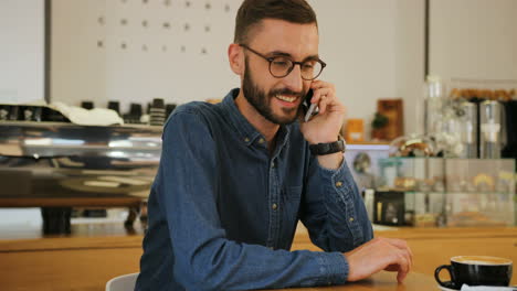 young caucasian man in glasses talking on smartphone while sitting at the table with cup of coffee in a coffee shop